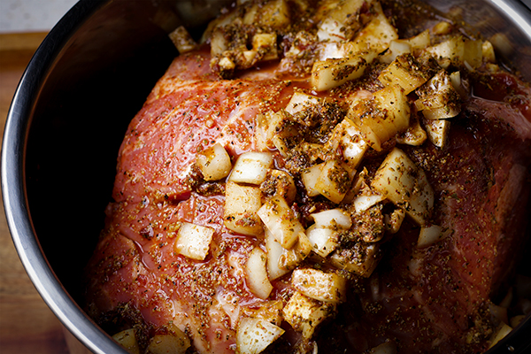 A pork shoulder roast in the bowl of an Instant Pot, covered in chopped onions and spices, ready to cook.