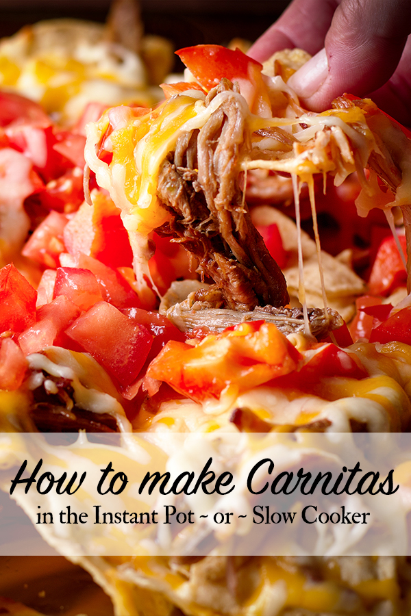 Someone lifting up a chip covered in cheese, carnitas, and fresh tomatoes from a tray of nachos.