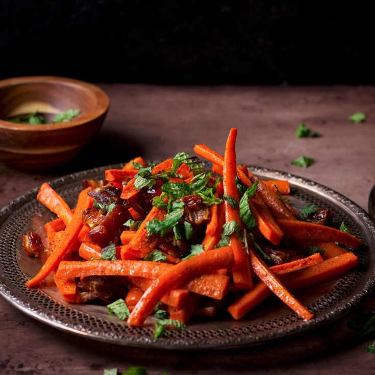 A silver platter piled high with roasted carrots and dates that have been sprinkled with fresh mint.