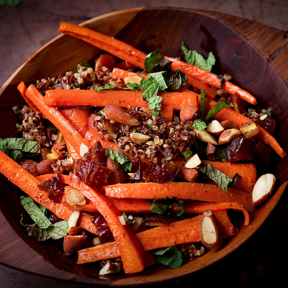 A wood bowl filled with cooked quinoa and roasted carrots and dates that have been sprinkled with roasted almonds and fresh mint.