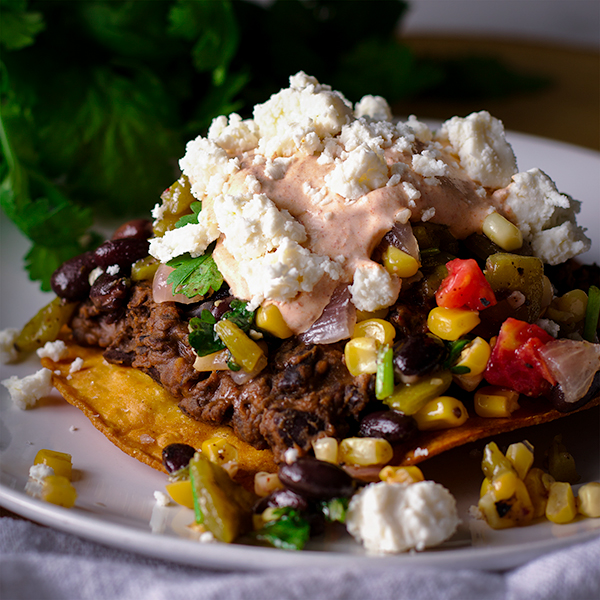 A Black Bean Tostada on a plate that's been topped with Corn Salsa, Enchilada Cream, and cheese.