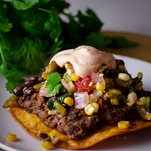 A Black Bean Tostada on a white plate that's been topped with Corn Salsa and Enchilada Cream.
