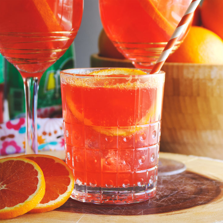 A wood tray with three glasses filled with Aperol Spritz. Fresh oranges surround the glasses.
