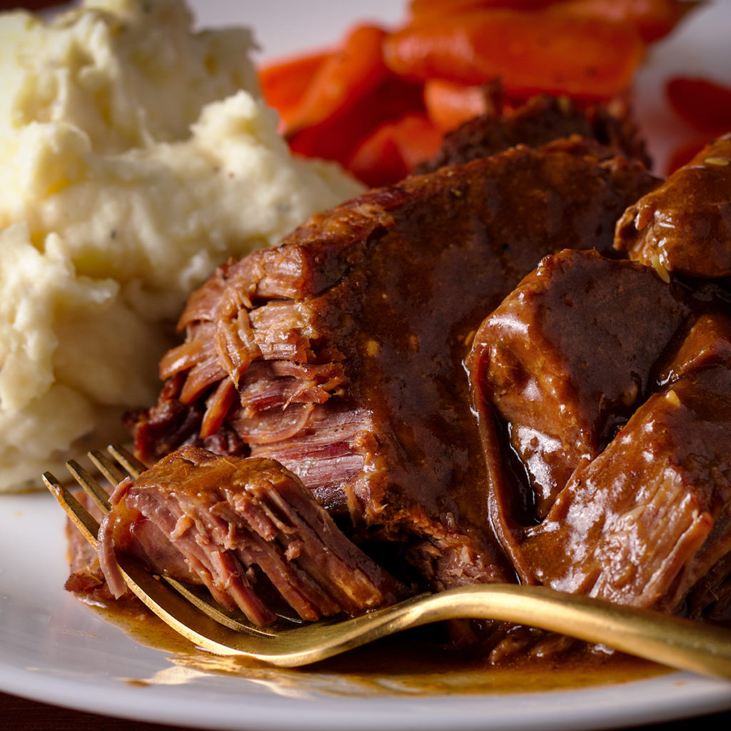 A white plate filled with cooked Tri-Tip Roast that's smothered in fennel and coriander gravy. A fork with a bite of meat rests on the plate.