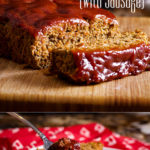 A partially sliced meatloaf made with sausage and covered with sweet and tangy glaze rests on a wood cutting board.