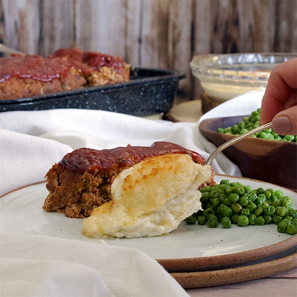 Spooning easy potatoes au gratin onto a plate with meatloaf and peas.