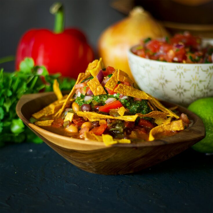 A wood bowl containing vegetarian chili topped with chimichurri, pico de gallo, and fried tortilla chips.