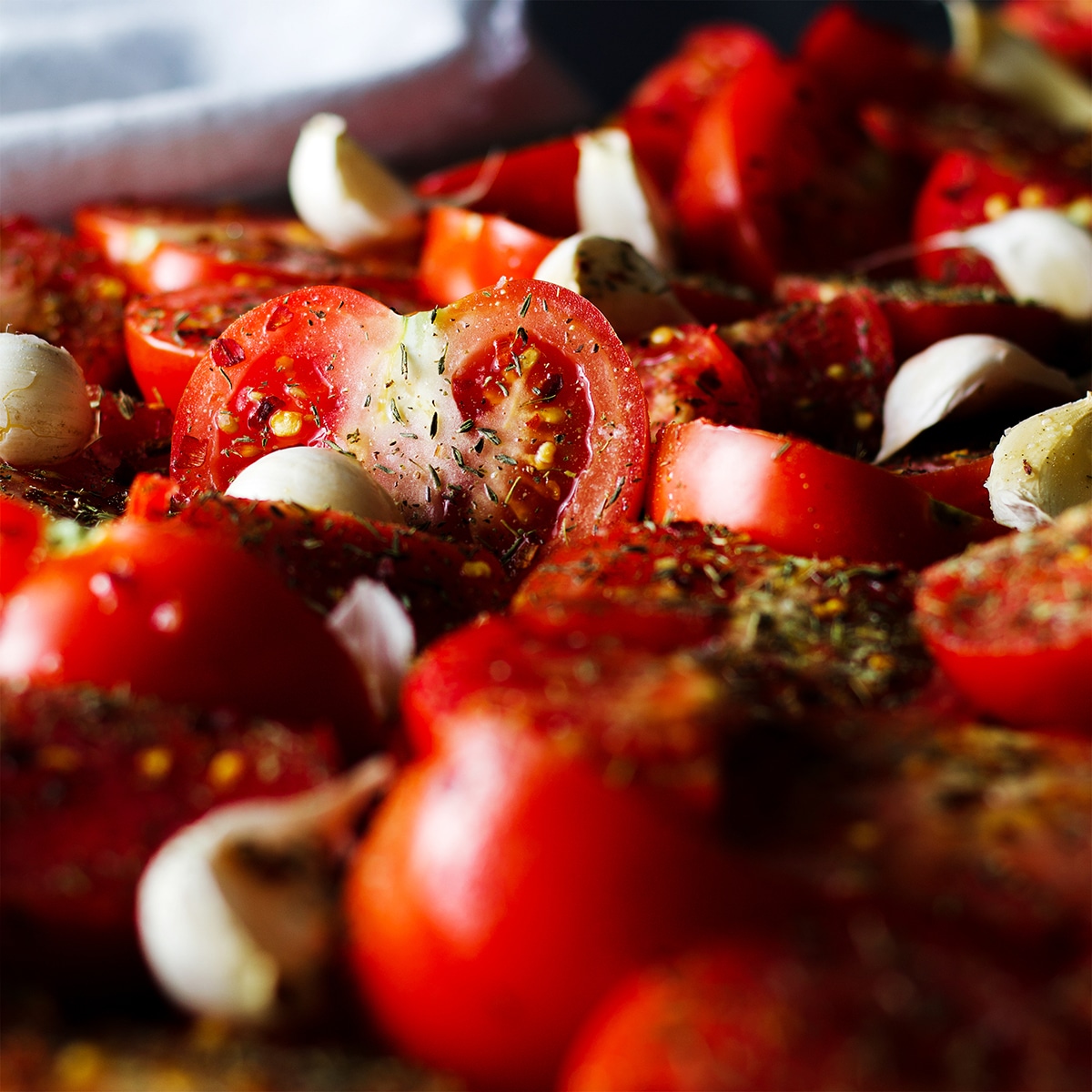 Ripe summer tomatoes that have been cut in half and spread out on a baking tray along with many cloves of garlic.