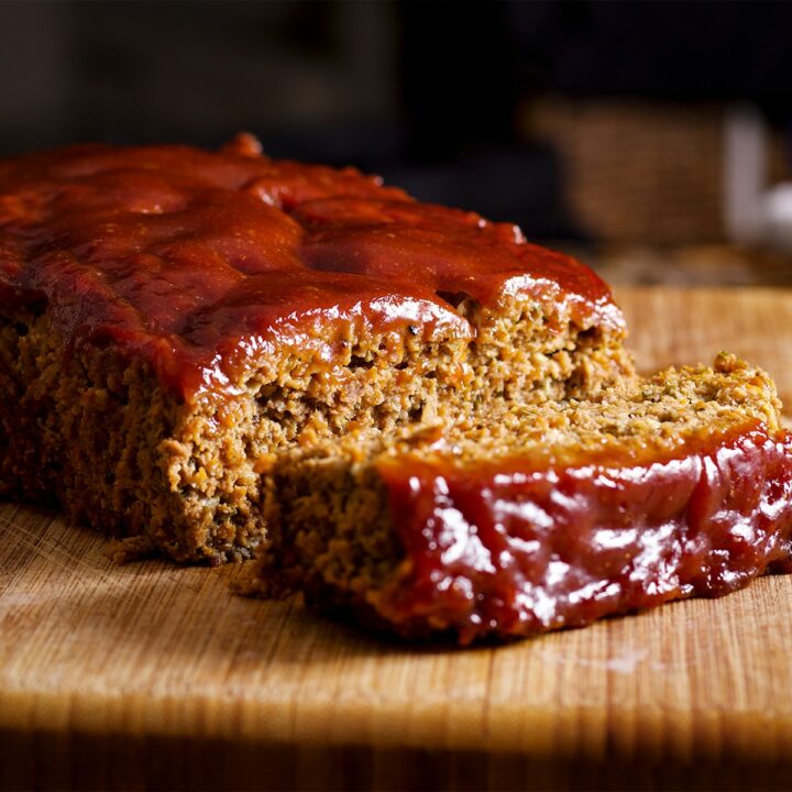 A partially sliced meatloaf made with sausage and covered with sweet and tangy glaze rests on a wood cutting board.