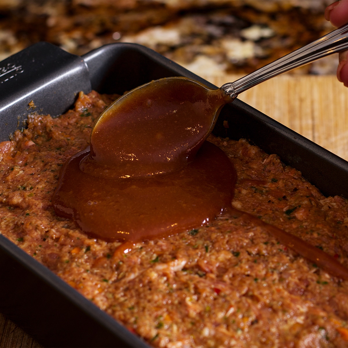 Someone using a spoon to spread glaze over raw meatloaf before baking.