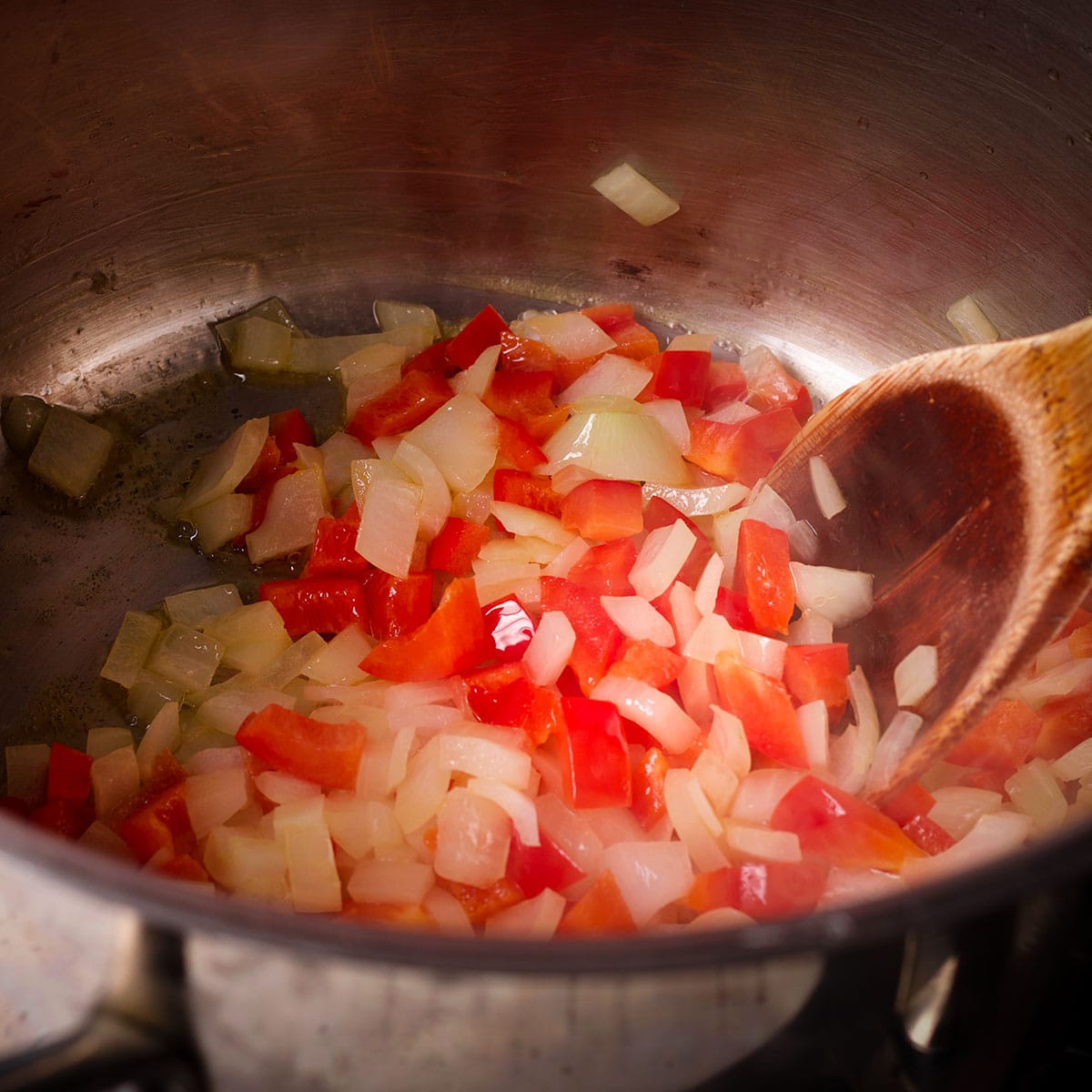 Using a wooden spoon to stir chopped onions and red bell peppers in a saucepan while they cook.