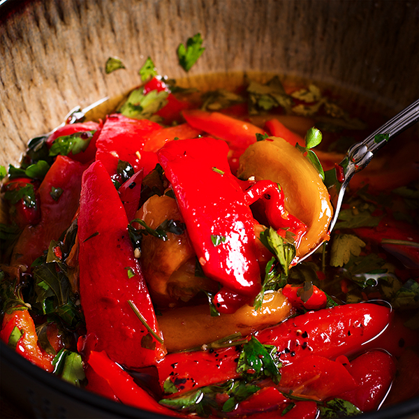 A bowl of marinated roasted bell peppers. Someone is using a silver spoon to lift a few peppers from the bowl.