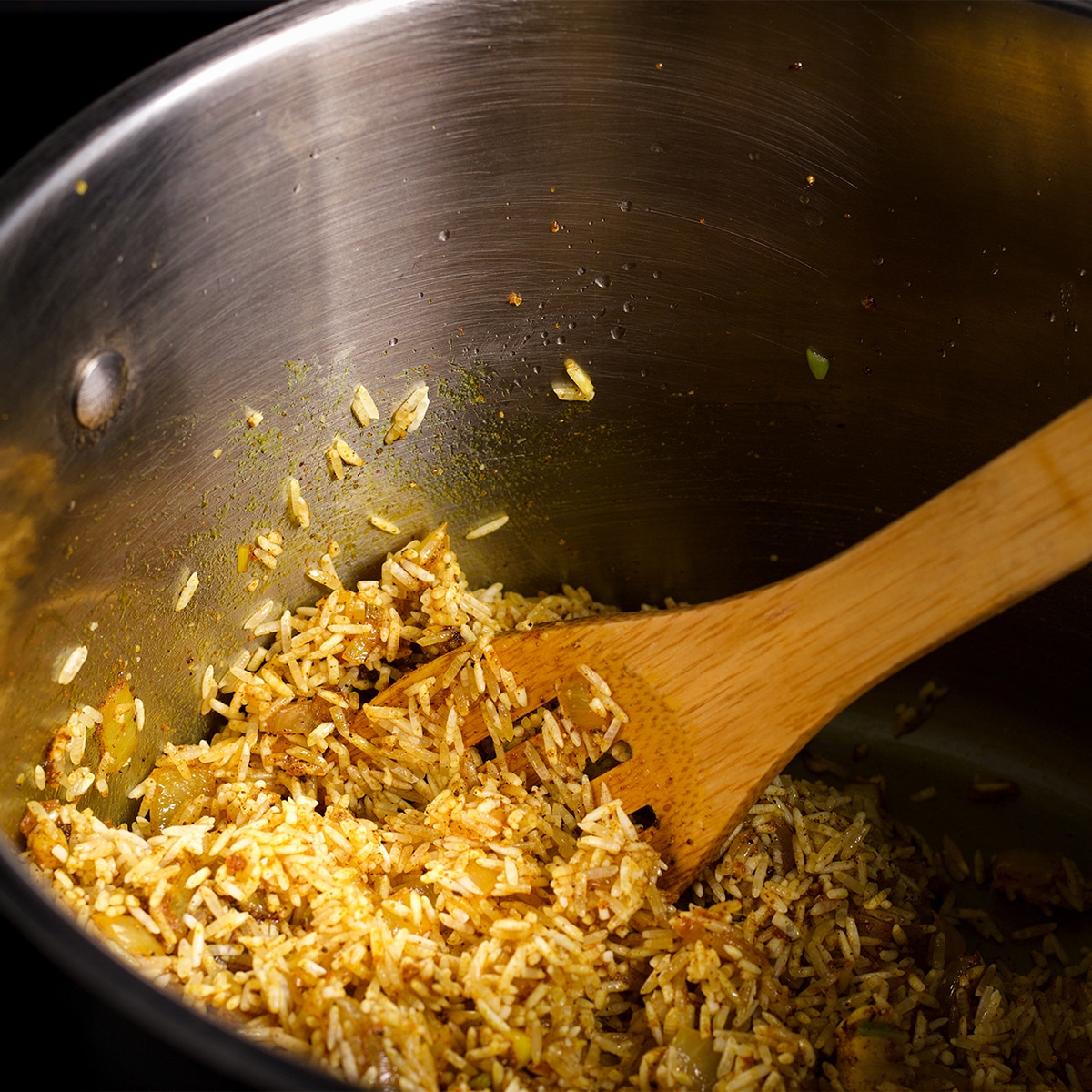 Using a wooden spoon to stir dry rice into onions and spices in a saucepan.