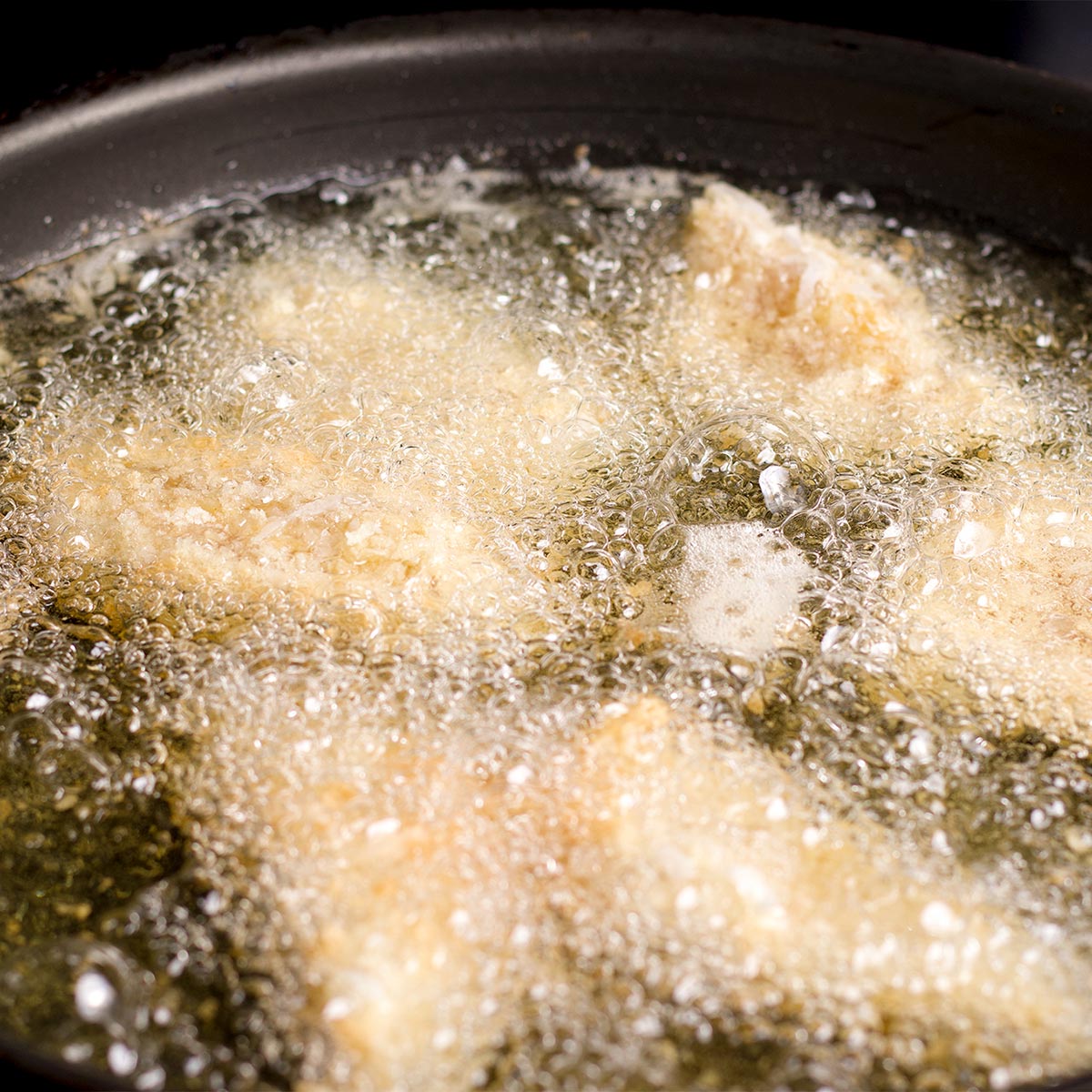 Pieces of coconut and panko breaded chicken frying in oil in a skillet set on a stovetop.