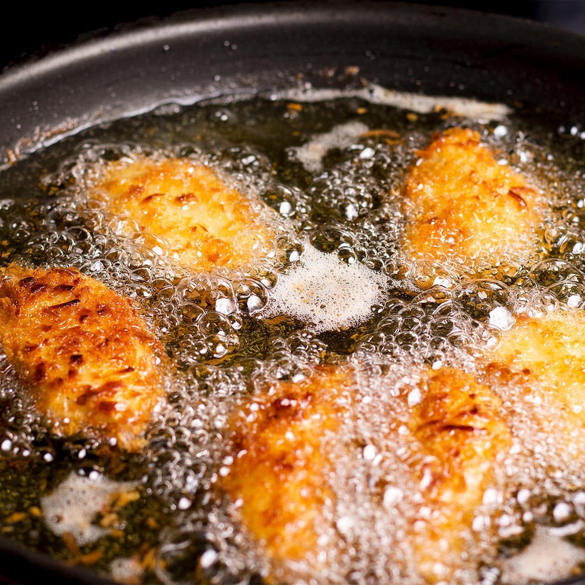 Pieces of coconut and panko breaded chicken frying in oil in a skillet set on a stovetop.