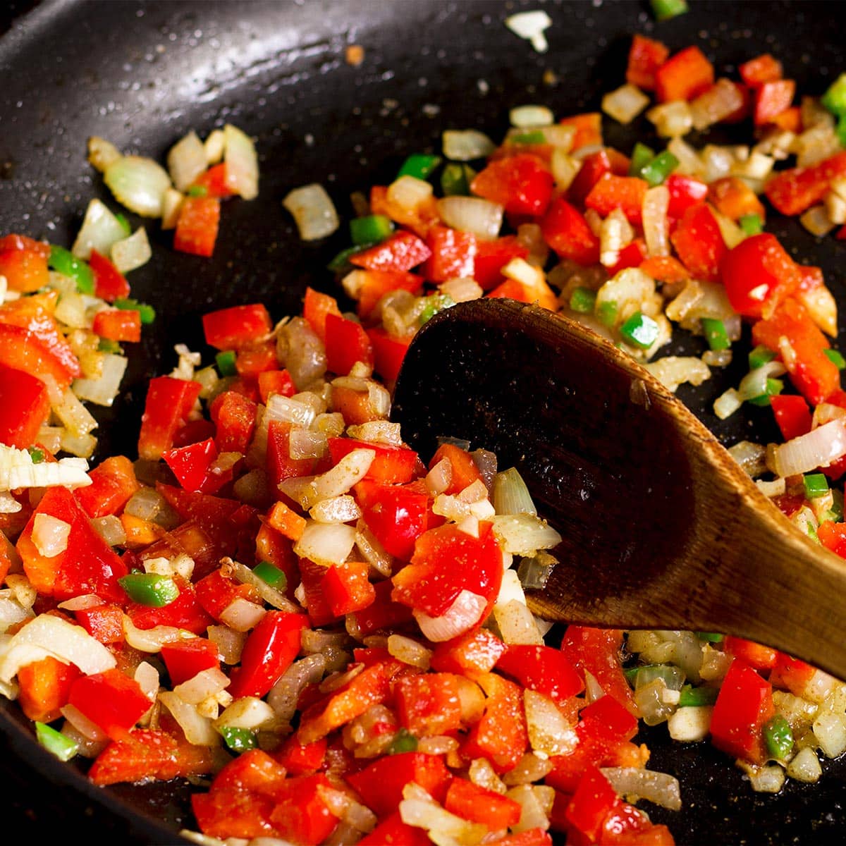 Using a wooden spoon to stir chopped red bell pepper, onion, garlic, and jalapeno in a skillet.
