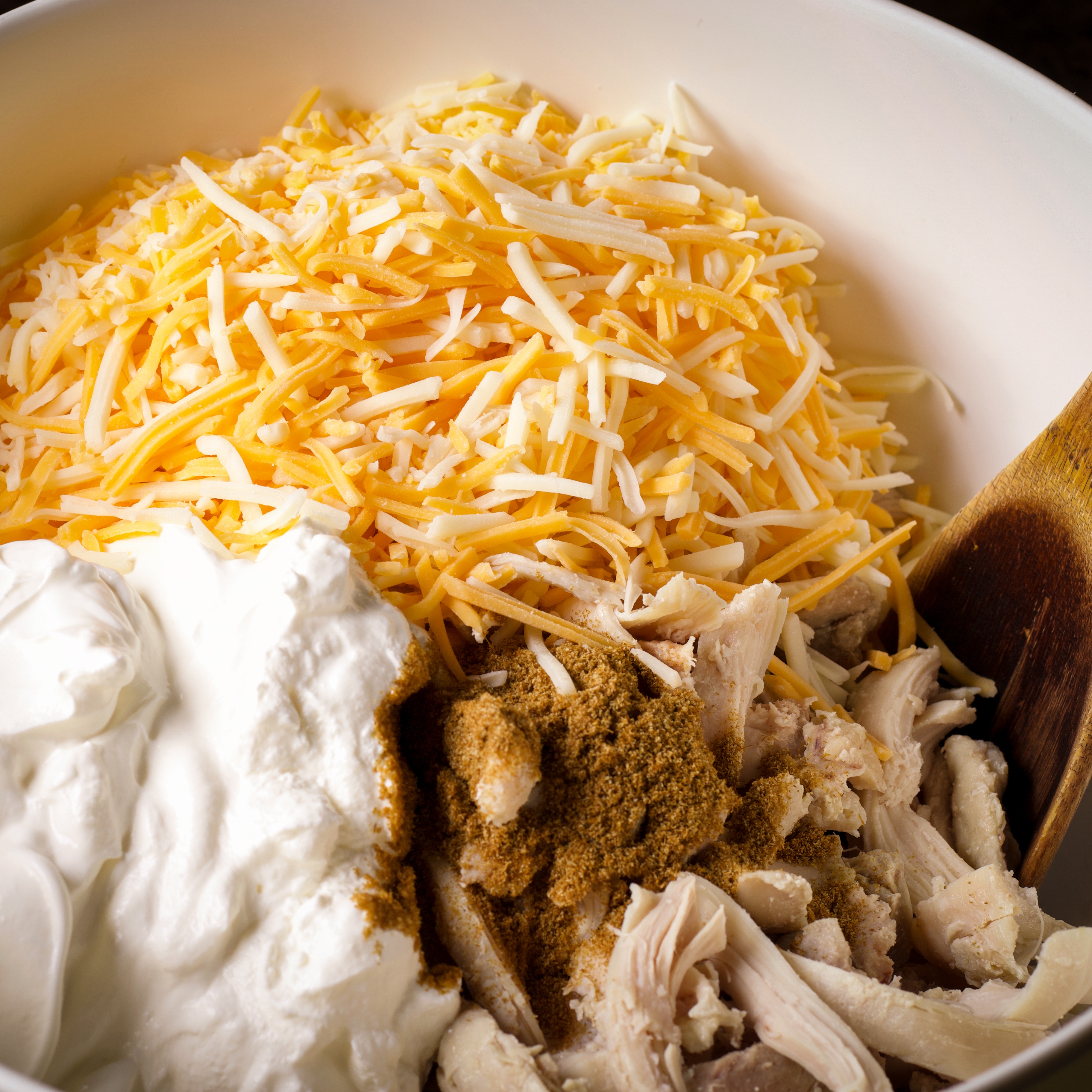 Adding grated cheese, cooked chicken, ground cumin, and sour cream to a bowl.