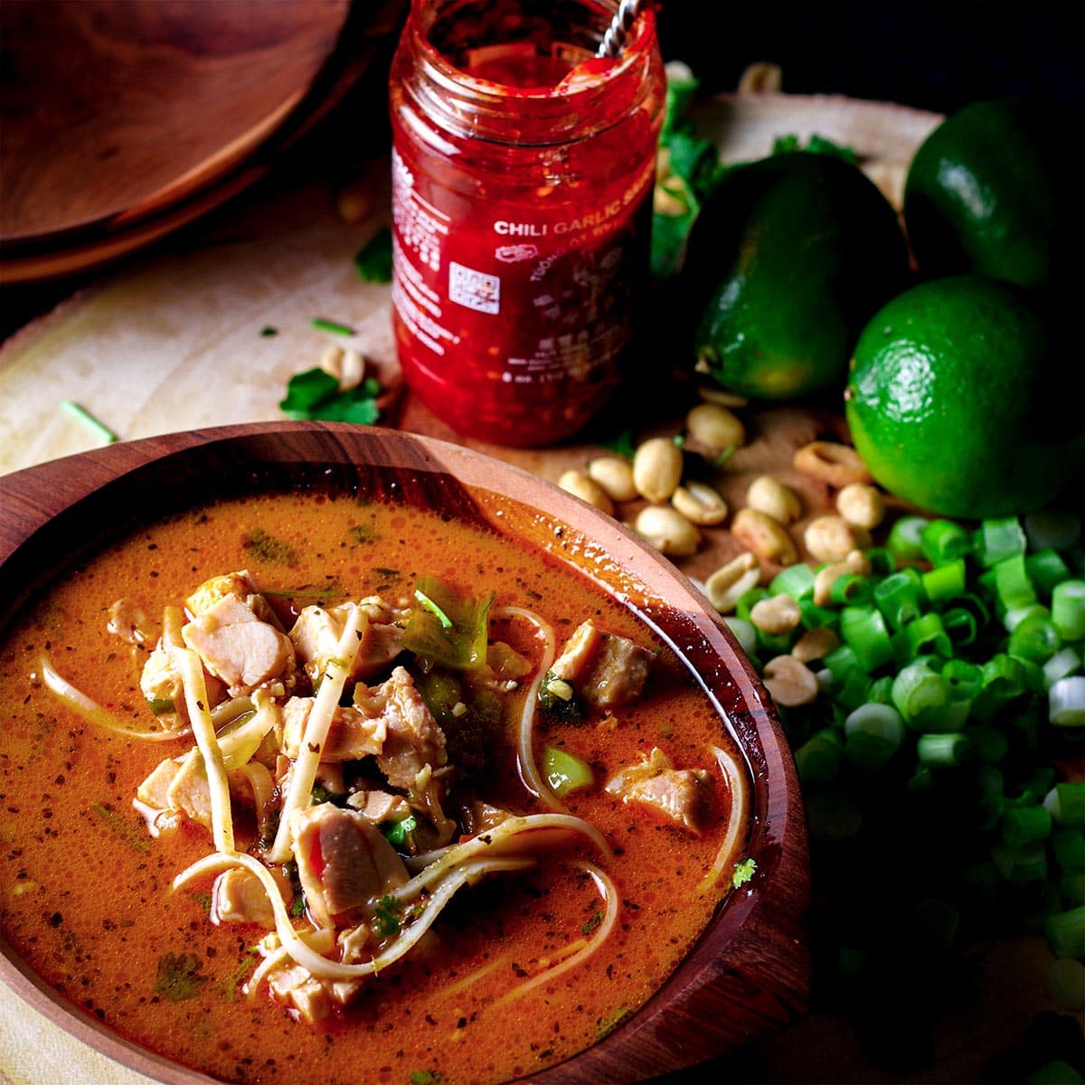A bowl of Thai chicken noodle soup on a table piled with chili garlic sauce, roasted peanuts, fresh limes, and cilantro.