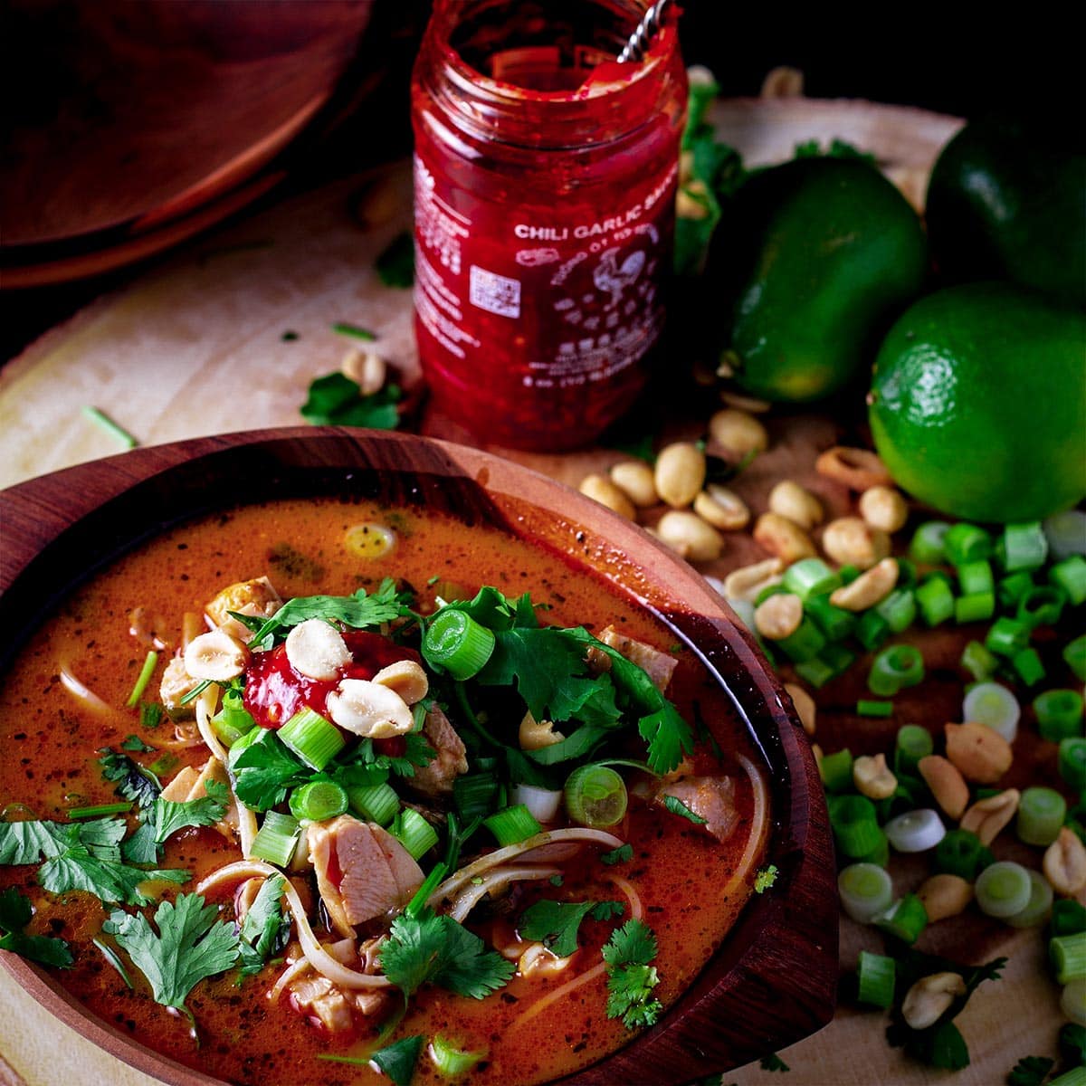 A bowl of Thai chicken noodle soup topped with chili garlic sauce, chopped cilantro, green onions, and roasted peanuts.