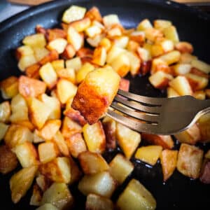 Someone using a fork to lifting a crispy fried potatoes from a skillet filled with cooking potatoes so you can see its golden edges.