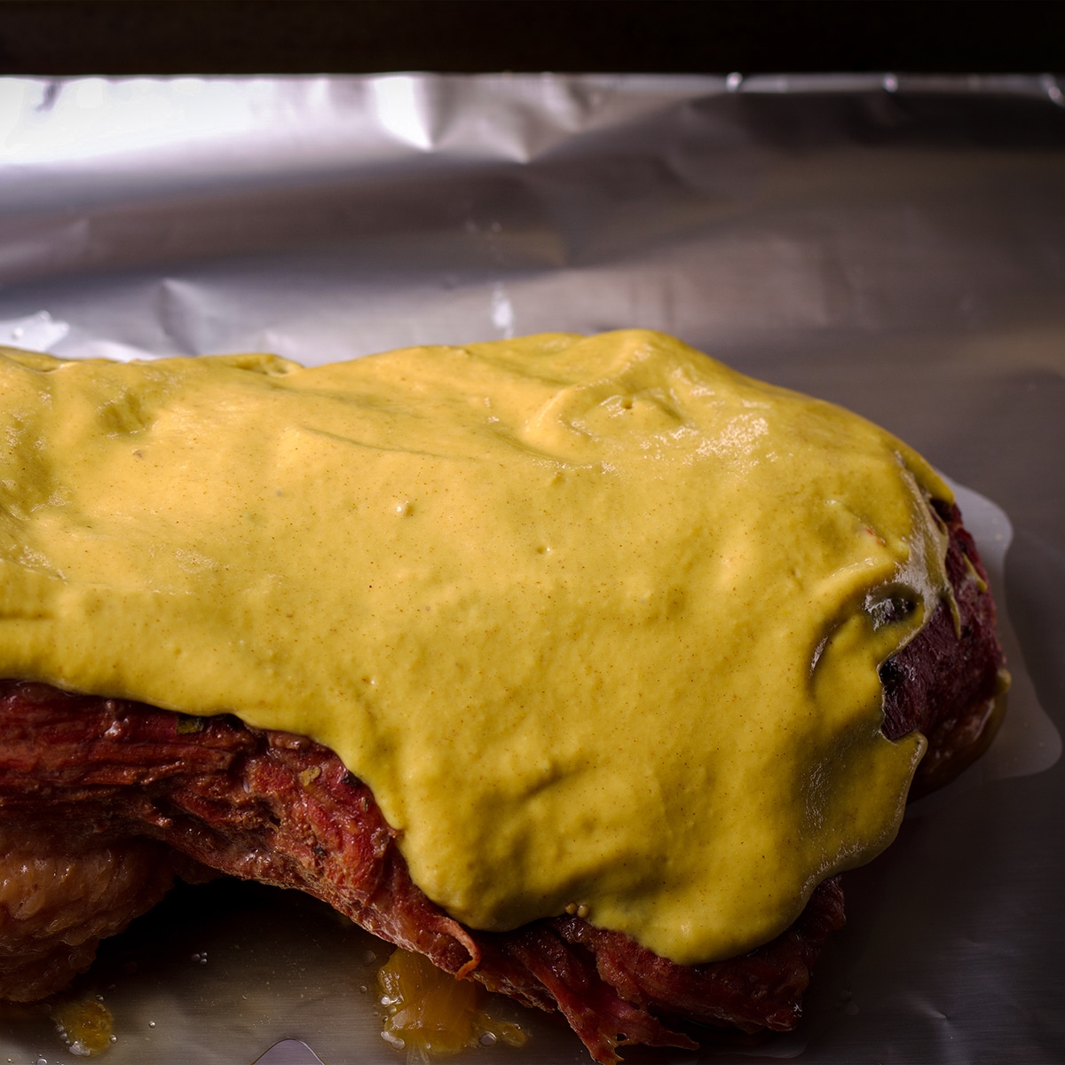 Someone spreading mustard sauce on a cooked corned beef on a baking sheet.