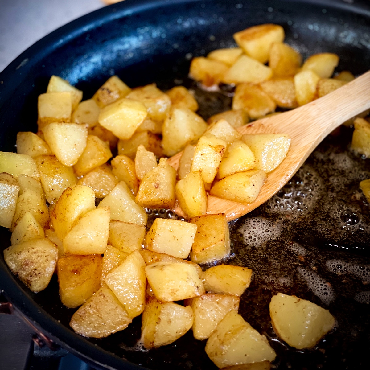 Someone using a wood spoon to stir potatoes in a hot skillet while they fry in butter and oil.