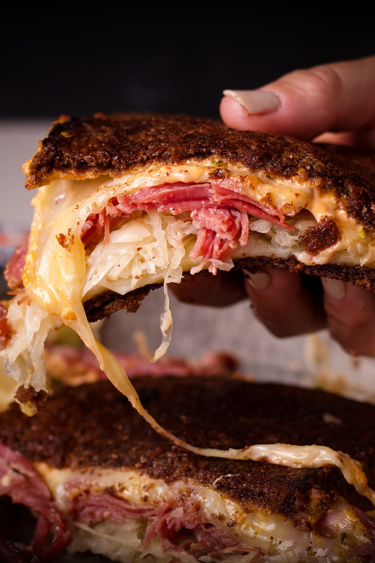 Someone lifting half of a Reuben sandwich from a plate with the cheese melting out of the sandwich.