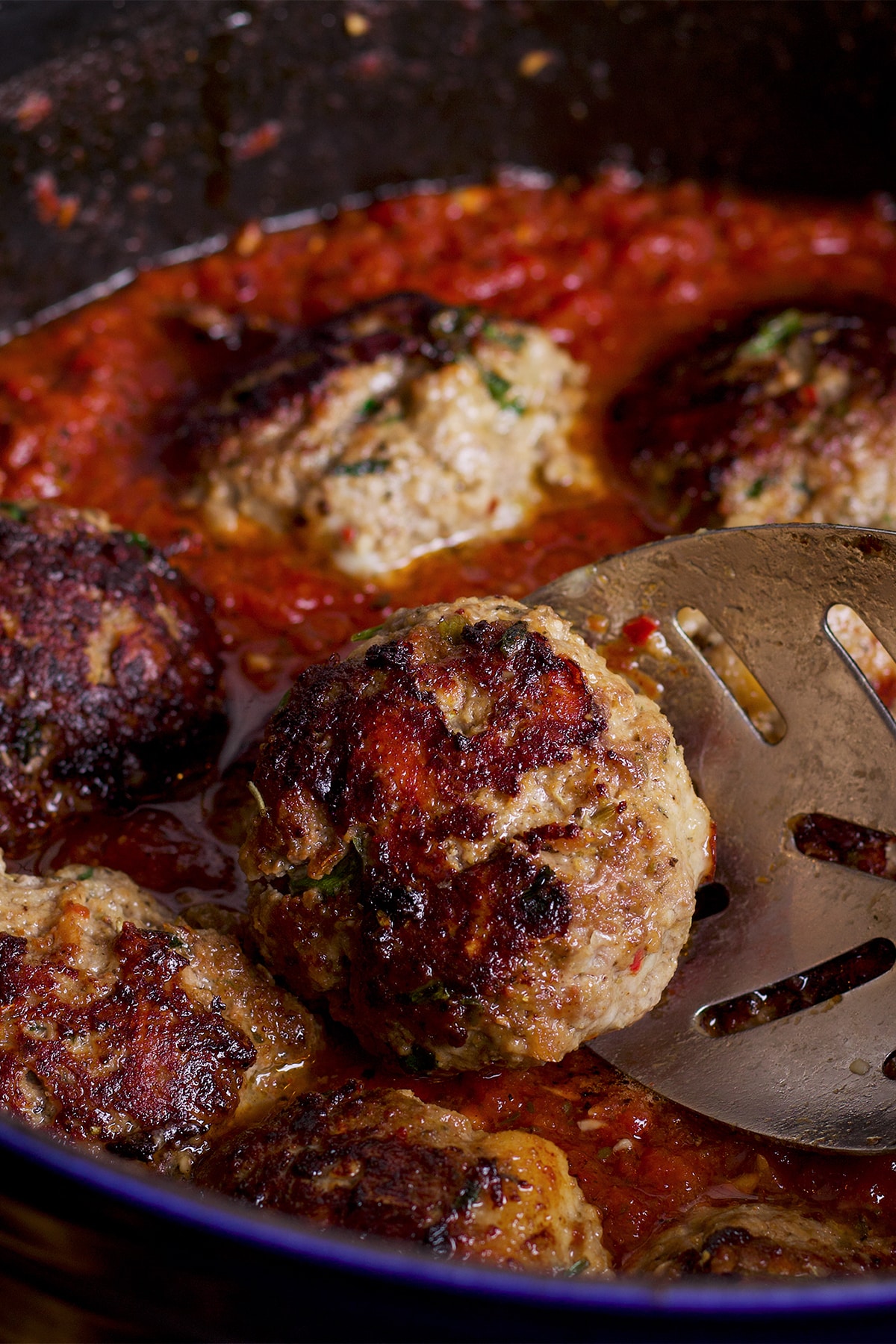 Someone using a spoon to nestle an Italian meatball into a pan filled with marinara.