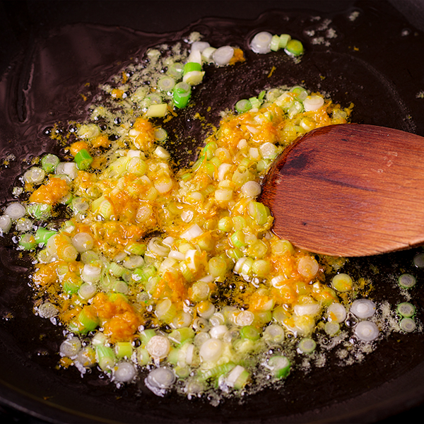 A wood spoon stirring grated ginger, orange zest, and scallions in a skillet with oil.