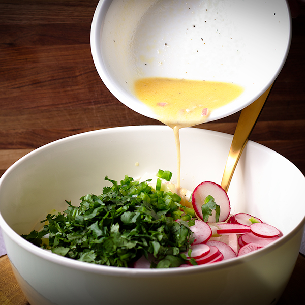 Pouring honey lime dressing over jicama, radishes, and cilantro in a white bowl.