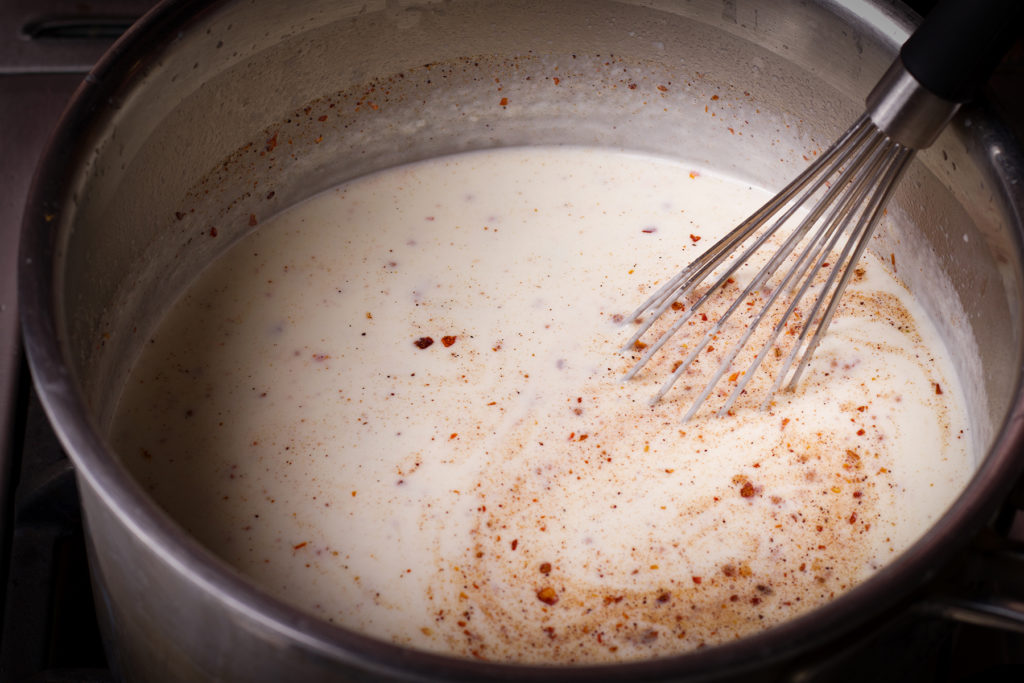 Whisking crushed red pepper flakes and ground nutmeg into a saucepan of béchamel sauce.