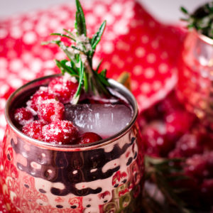 Two Cranberry Moscow Mule cocktails in copper mugs garnished with sugared cranberries and fresh rosemary.
