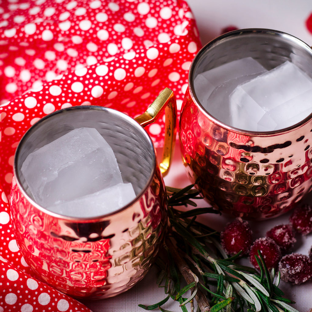 Two copper Moscow Mule mugs filled with ice sitting on a white table with a red and white polkadot napkin next to them.