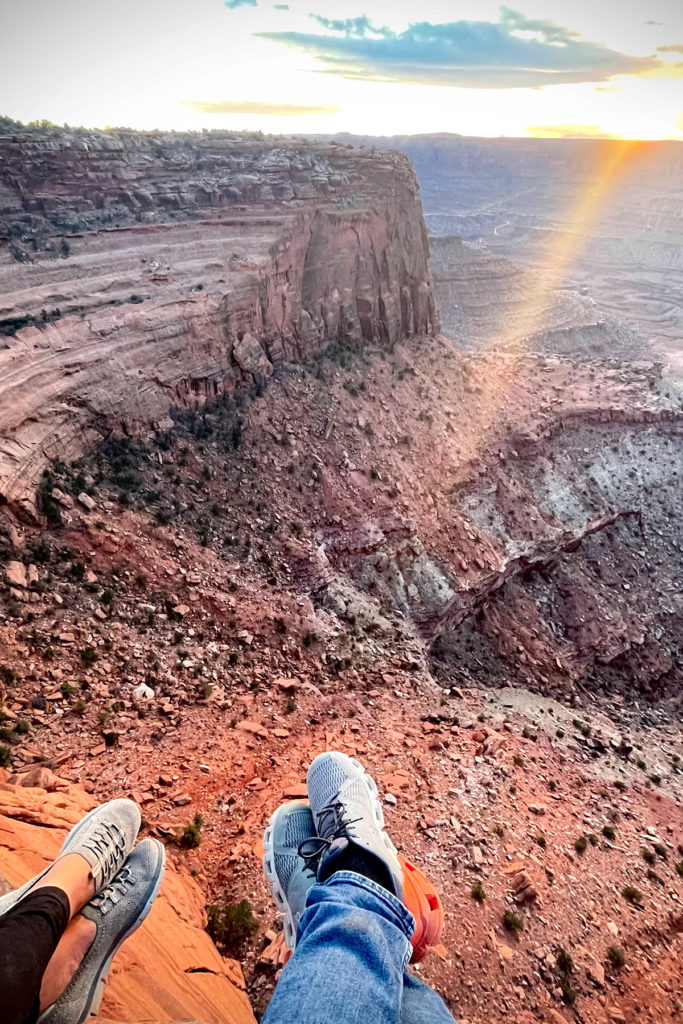 Two people sitting on the ground at Dead Horse Point state campground with their feet dangling off the edge of the canyon's edge.