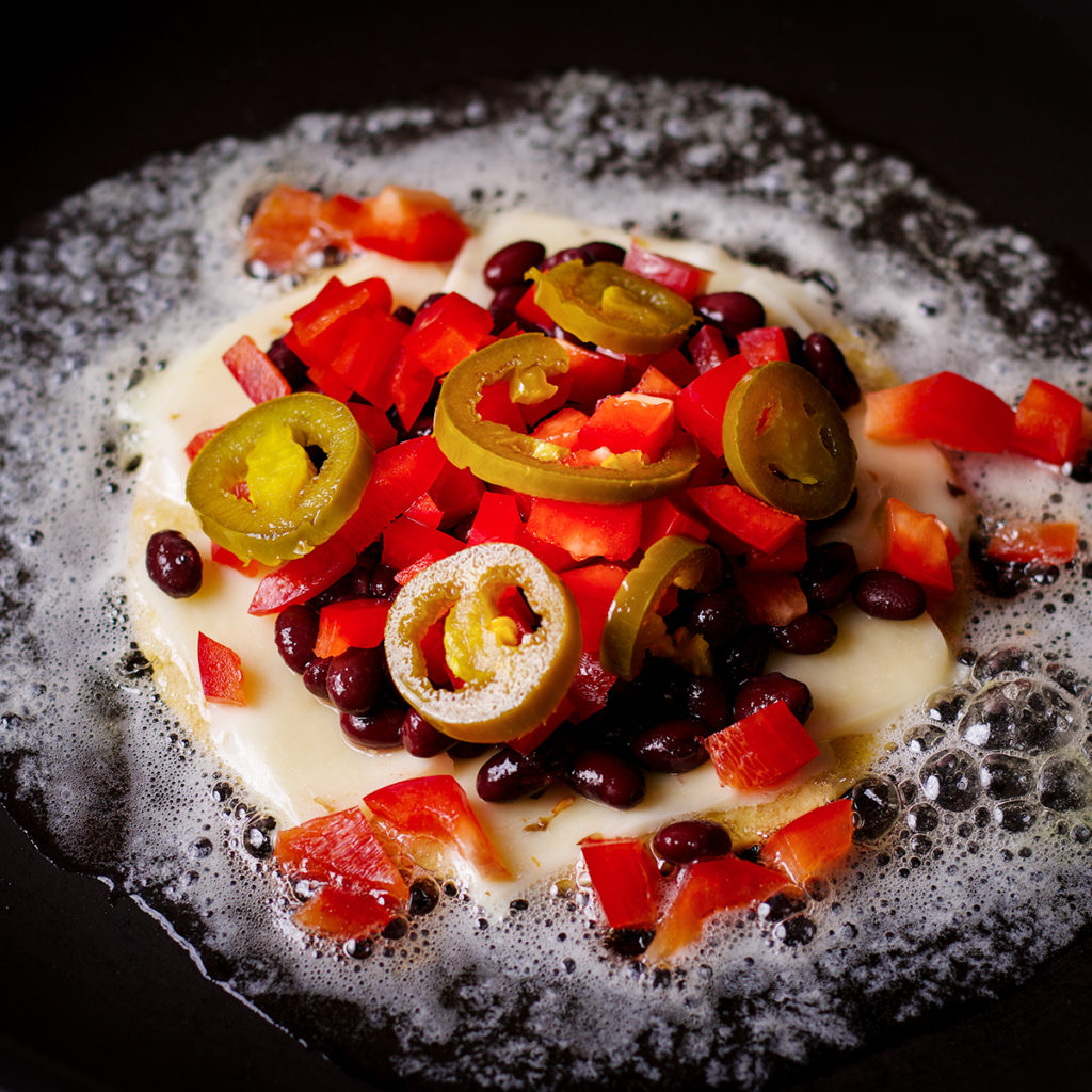 A corn tortilla in a frying pan, topped with slices of pepper jack cheese, black beans, chopped red bell peppers, and pickled jalapeño slices.