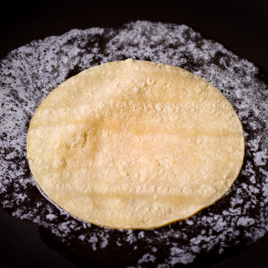 A corn tortilla cooking in butter in a frying pan.