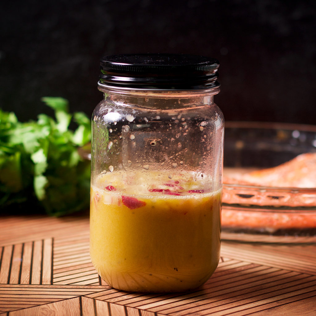 A jar of citrus vinaigrette on a wood table with baked salmon and a bunch of cilantro in the background.