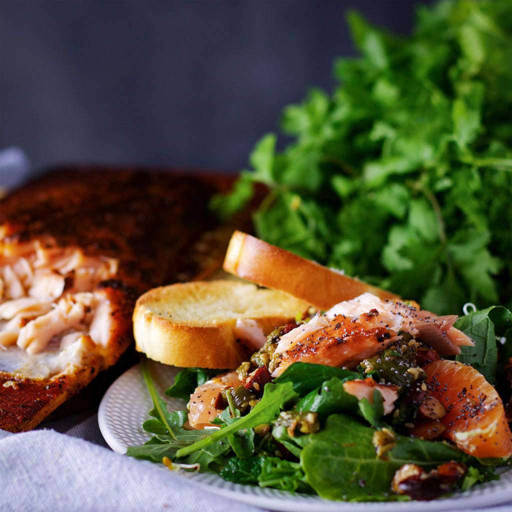 A plate piled high with salmon salad with citrus vinaigrette and almond salsa and a couple of pieces of toasted bread. A large piece of baked salmon is in the background.