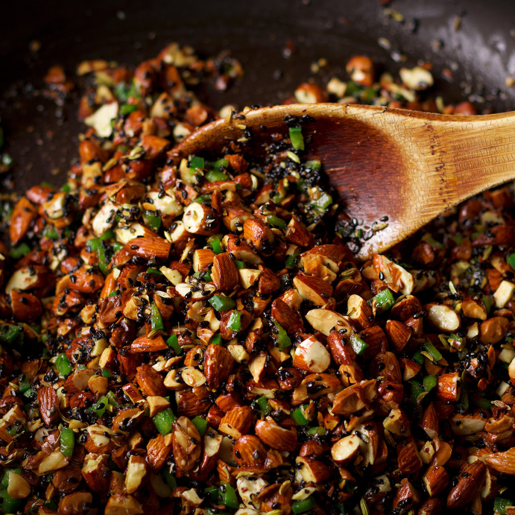 Someone using a wooden spoon to stir a skillet full of almond salsa with green chilies.