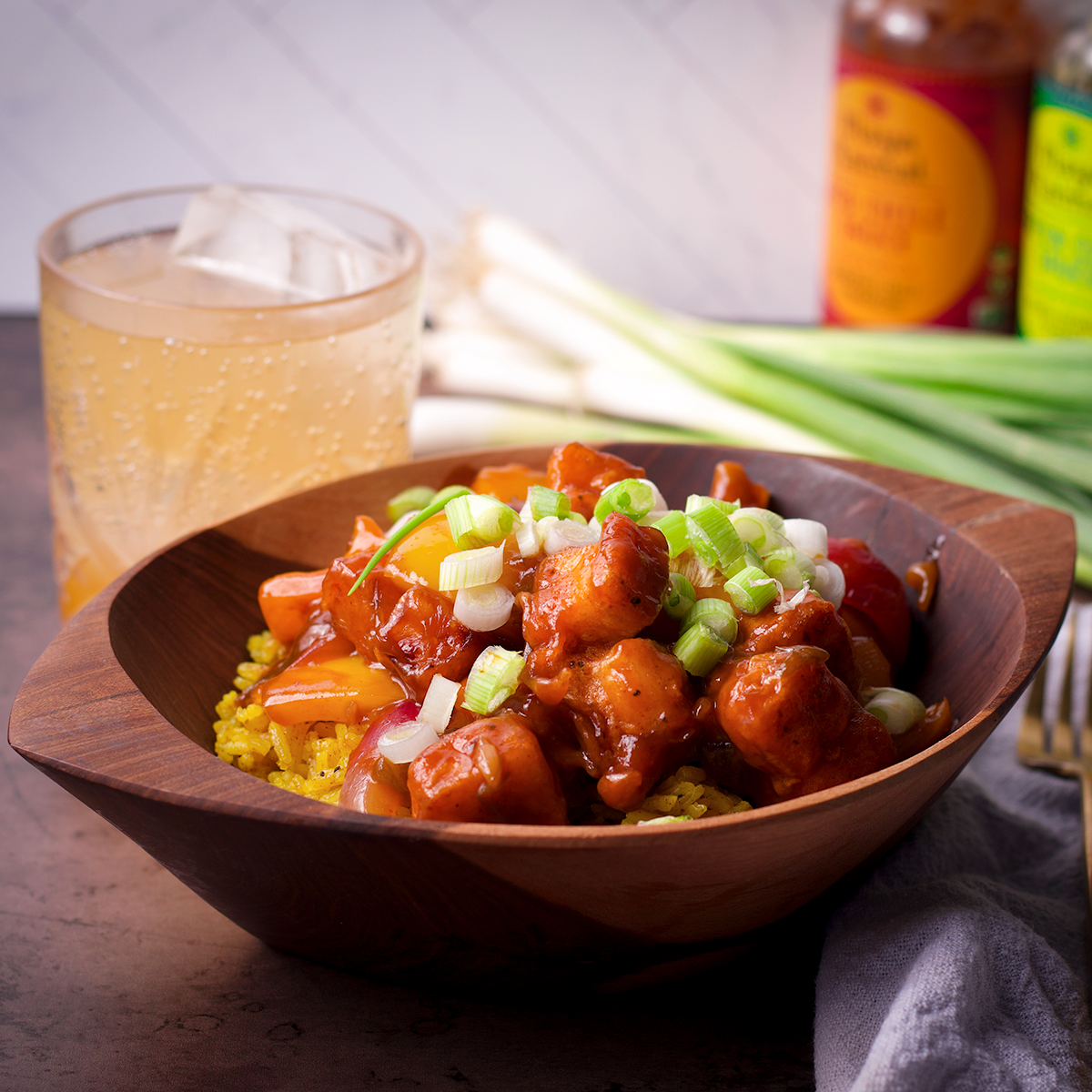 A wood bowl filled with rice and chilli paneer. In the background is a drinking glass filled with soda, green onions, and a couple of different kinds of hot sauce.