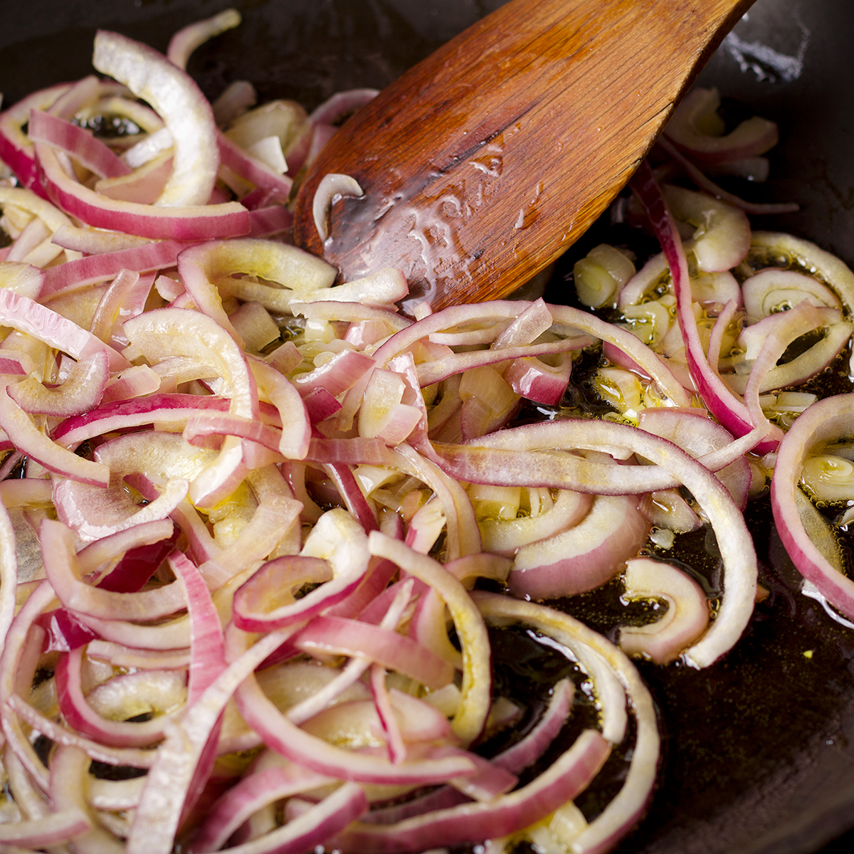 Someone using a wood spoon to stir onions cooking in a skillet.