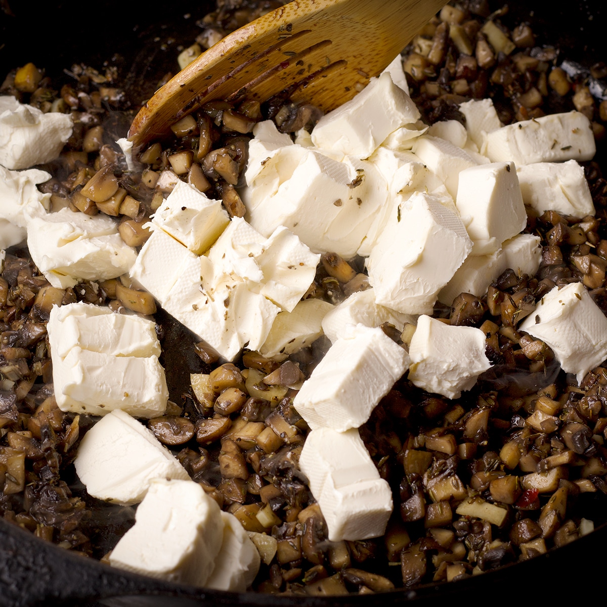 Cubes of cream cheese in a hot skillet that's also filled with diced mushrooms.
