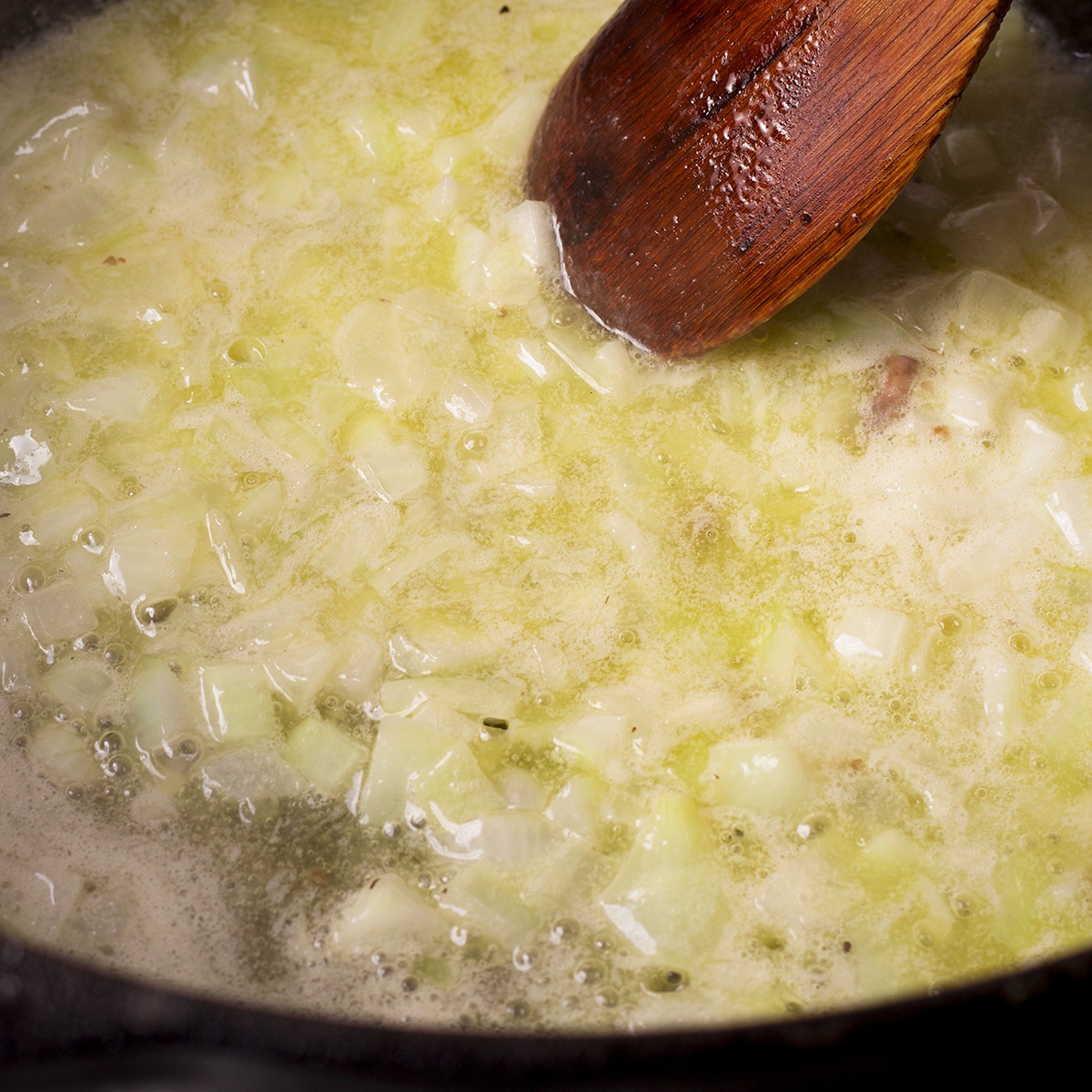 Someone using a wooden spoon to stir onions simmering in butter.