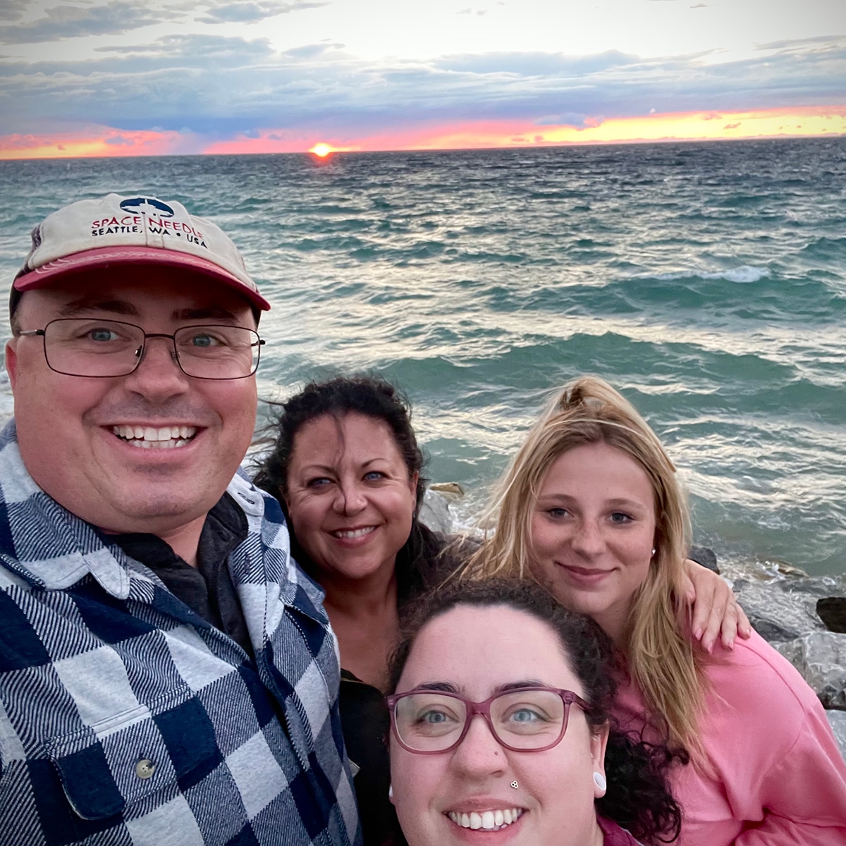 Steve and I with our daughter and niece standing on a Lake Michigan beach in Petoskey with a brilliant sunset over the water behind us.