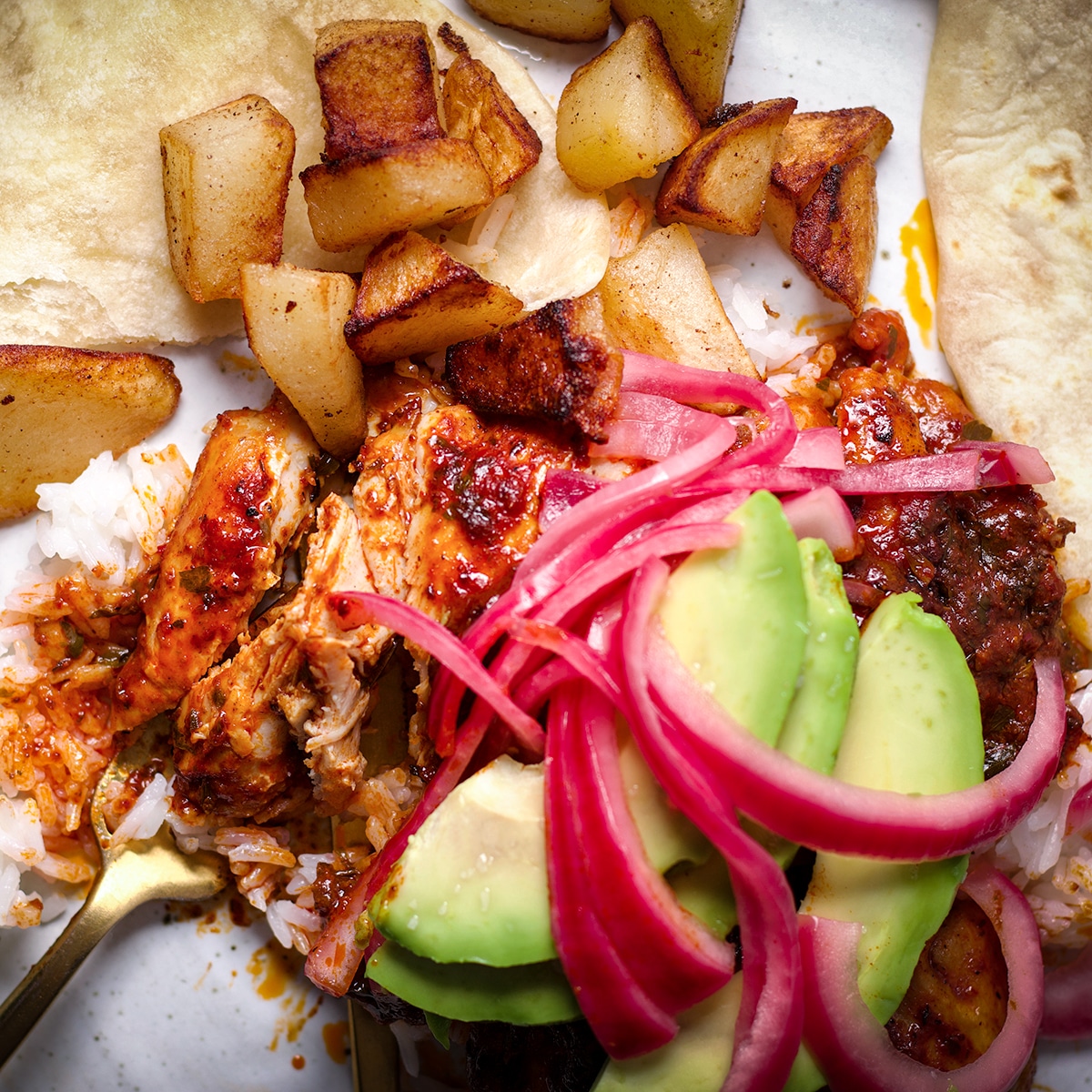 A photo looking down onto a plate of Mexican Adobo Chicken served with rice, crispy fried potatoes and a flour tortilla. A fork has cut into the chicken so you can see the tender, juicy inside of the meat.