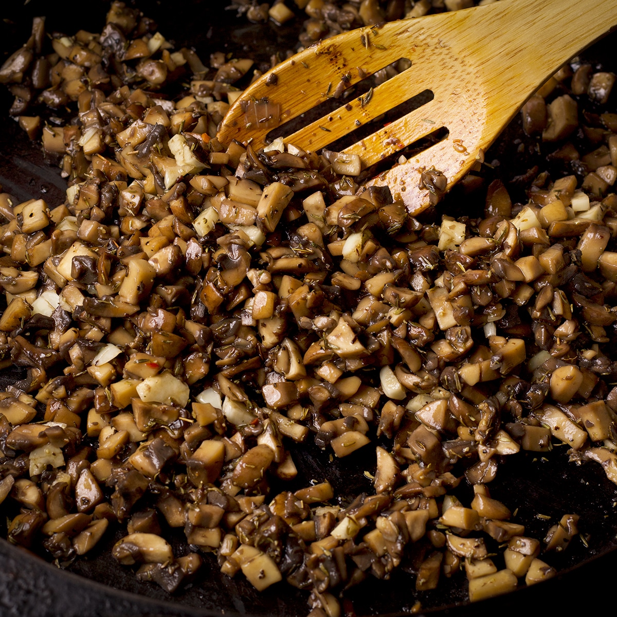 Someone using a wooden spoon to stir diced mushrooms as they cook in a cast iron skillet,