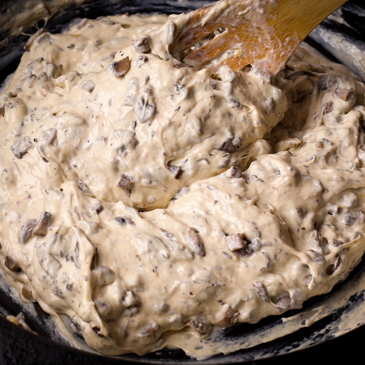Someone using a wooden spoon to stir cream cheese into mushroom dip.