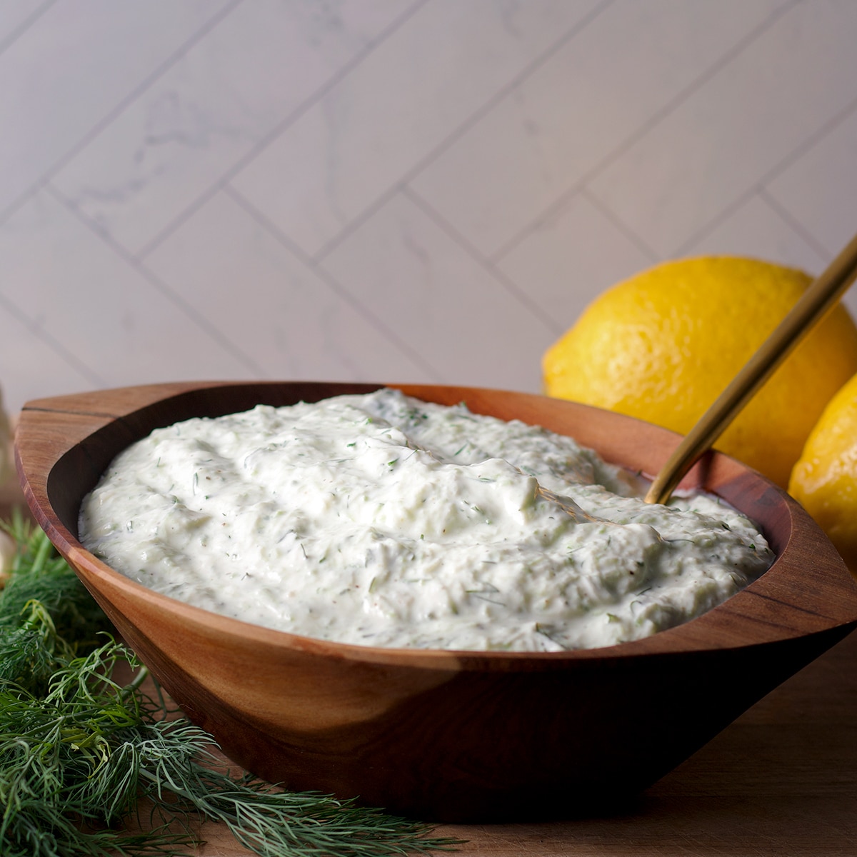 A wood bowl filled with tzatziki sauce with fresh dill and lemons on a cutting board next to the bowl.