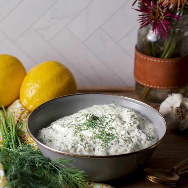 A blue bowl containing vegan tzatziki sauce on a wood board surrounded by fresh dill and lemons.