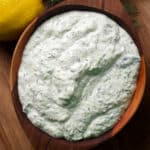A bowl containing tzatziki sauce rests on a wood cutting board surrounded by fresh dill and lemons.